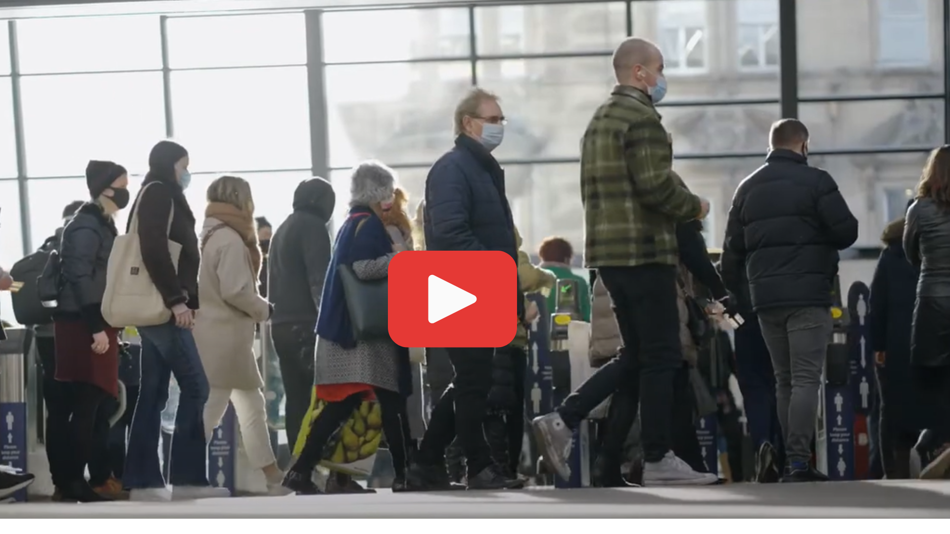 Passengers in a railway station. The image has a play button on it to indicate a link to YouTube. 