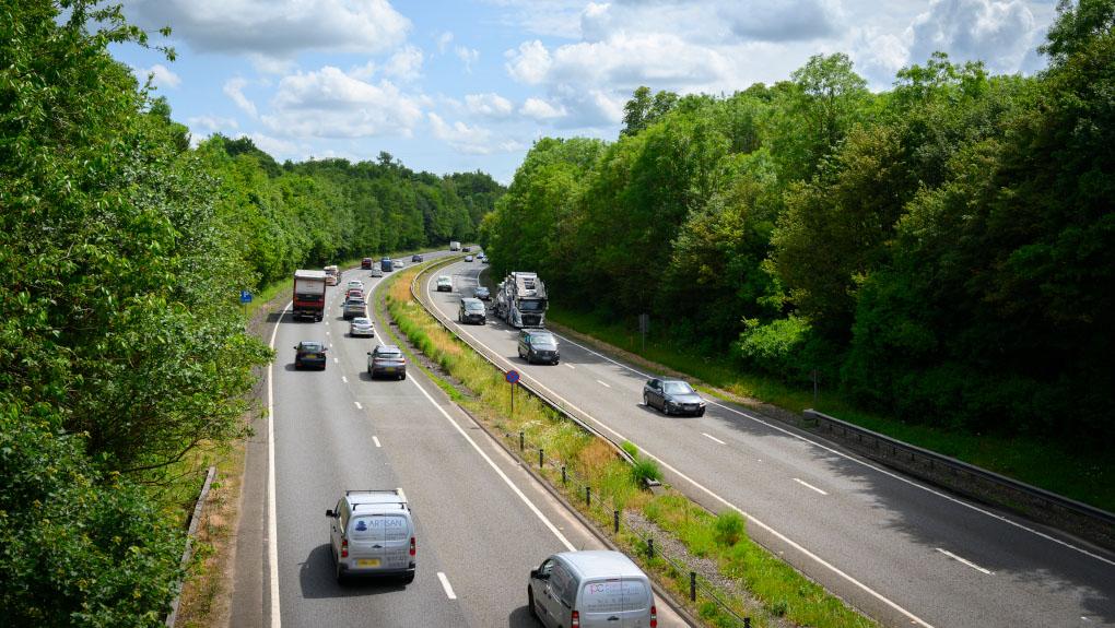 A dual carriageway in England with vehicles driving on it. Tall green trees line the side of the dual carriageway.